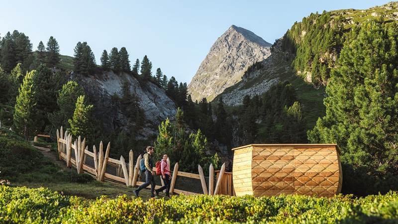 Zwei Wanderer gehen einen Hügel hinab, umgeben von Wiesen, Bäumen und Bergspitzen.