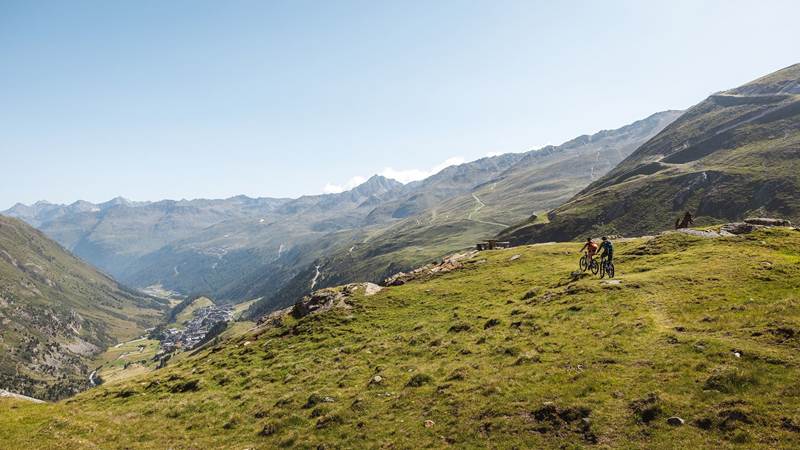 Zwei Radfahrer auf auf einem Bergpfad umgeben von Wiesen und Bergen.