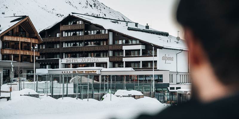The Hotel Alpina from the outside in the snow, in the foreground the silhouette of a man.
